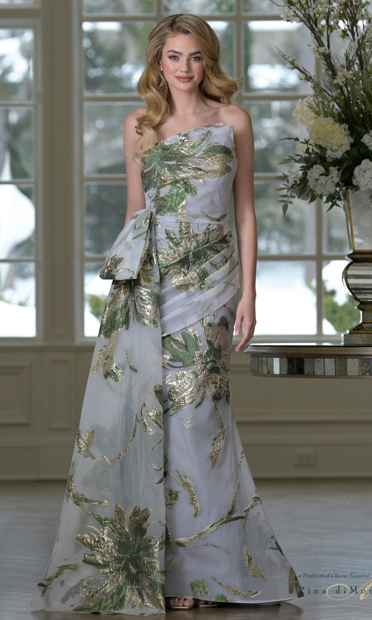 A woman stands indoors wearing the One-Shoulder Formal Gown: Rina Di Montella RD3233, a floor-length organza jacquard dress with metallic green and gold floral print, featuring a one-shoulder design and a large side bow.